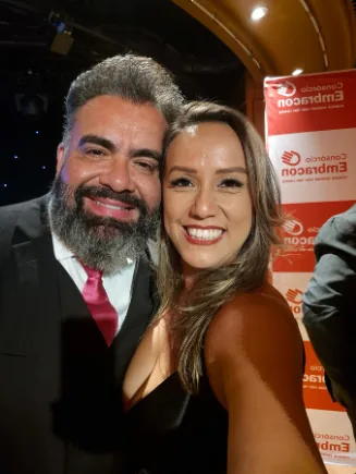 Ariane e William, sorrindo lado a lado em um evento, com painel da Embracon ao fundo